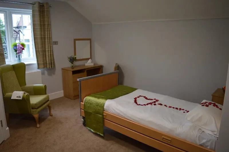 A bedroom with light wood bed frame and draw with white sheets, red petals and an olive green throw on the bed.