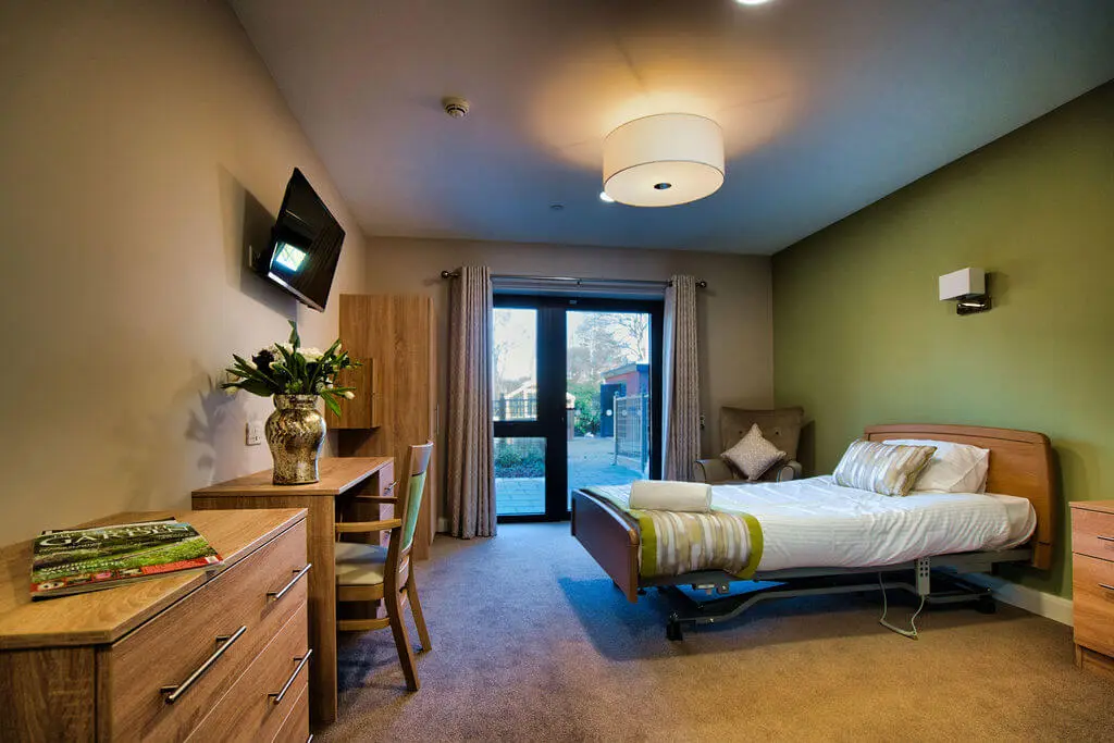 A bedroom with olive green feature wall, a bed, a table, a chest of drawers, a tv, a floor to ceiling window and some flowers.