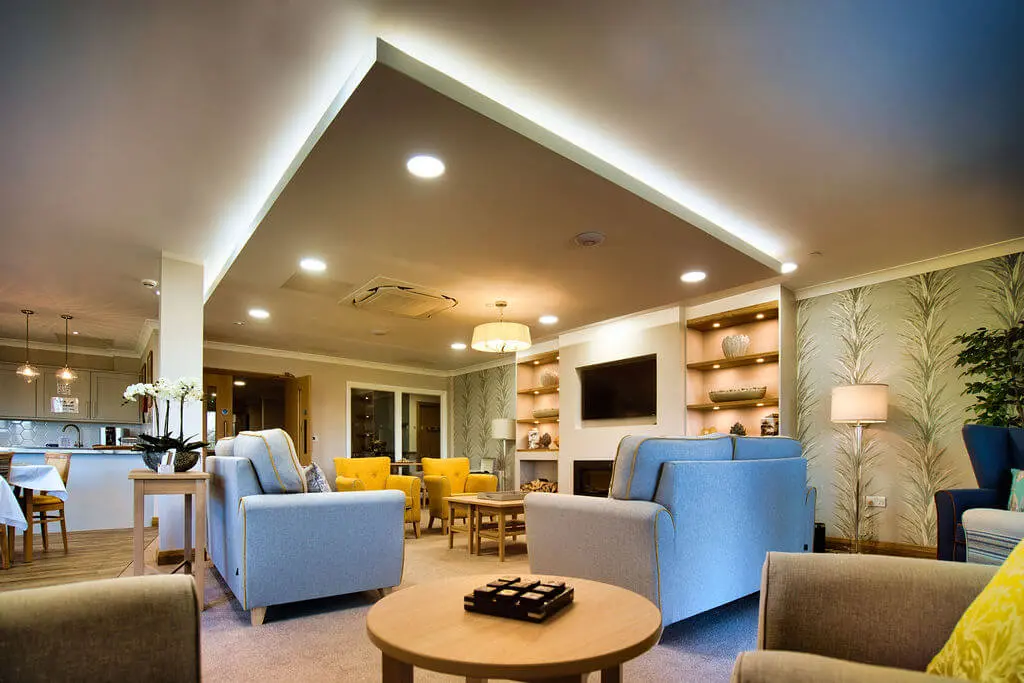 Tables, sofas and arm chairs in a communal lounge with TV mounted on the wall.