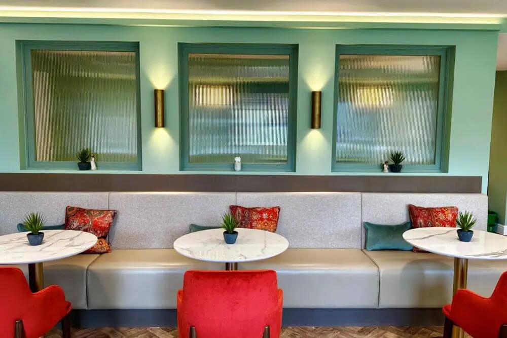 A grey booth bench with cushions, a round table and a red chair opposite the bench.