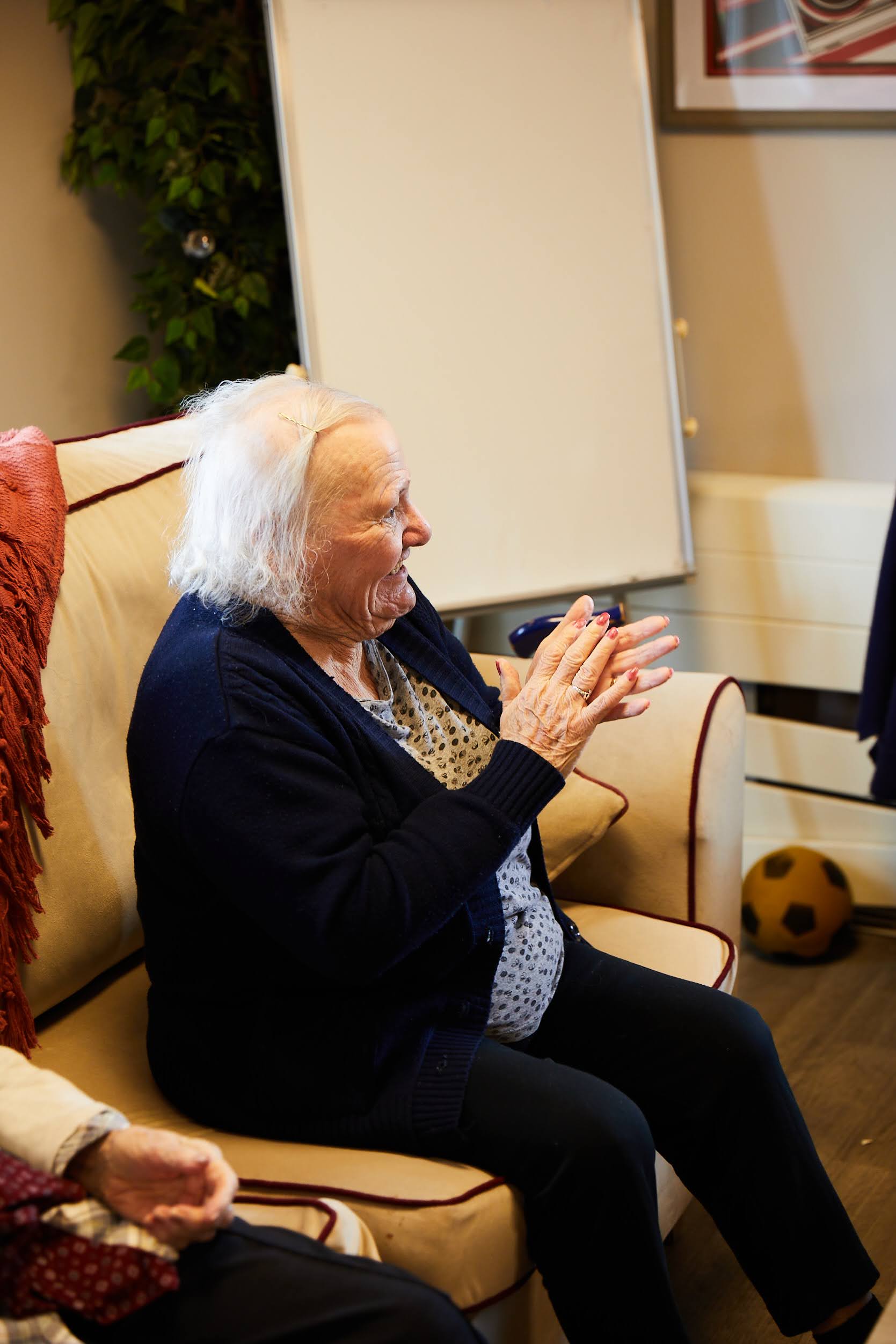 A lady sat down on a sofa clapping in the lounge at Brampton Lodge Day Centre