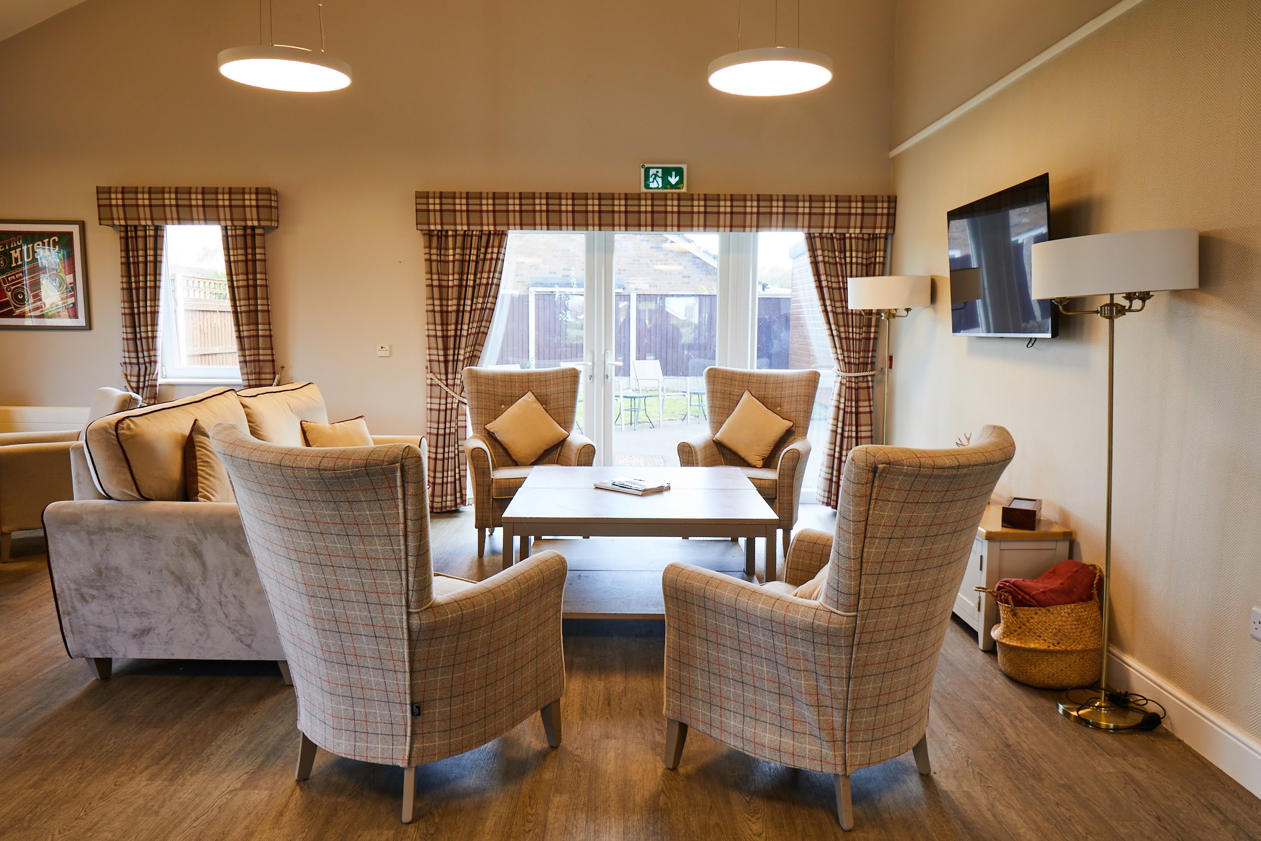 Arm chairs with cushions, a table, a TV mounted on the wall and a view out of large windows behind them in the Brampton Lodge Day Centre lounge.
