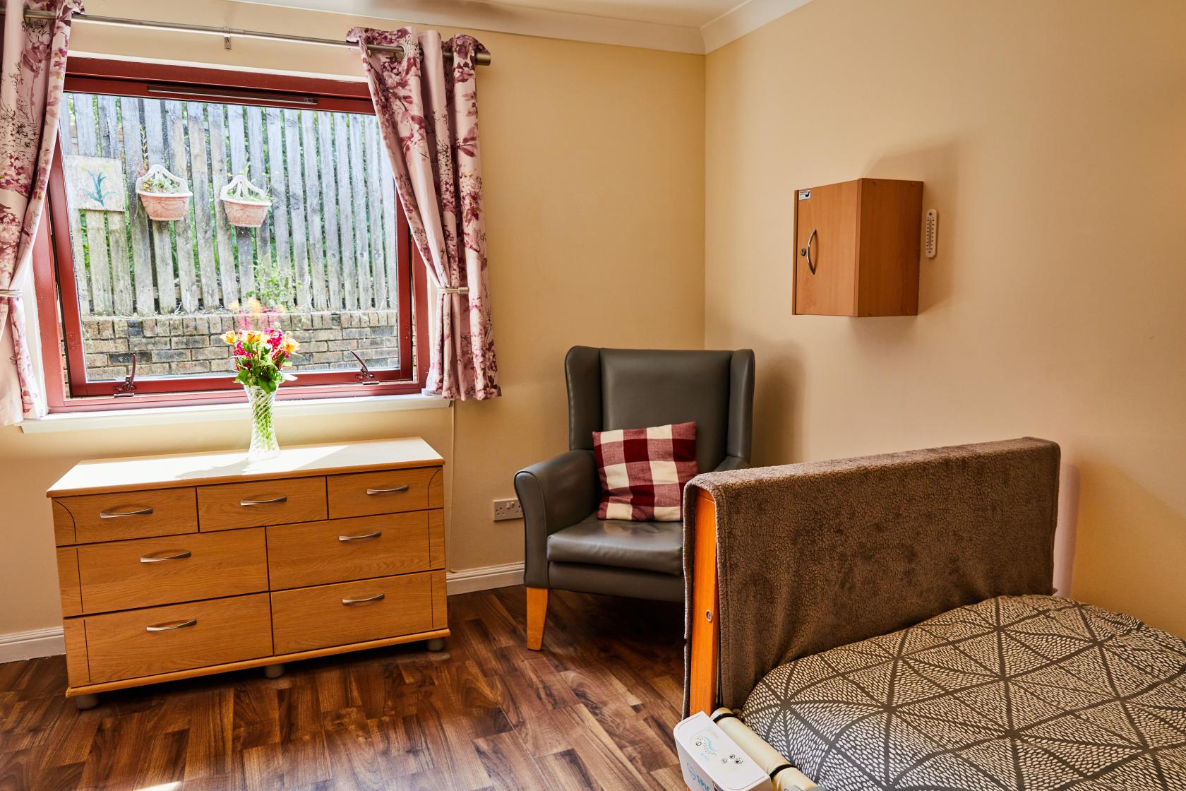 A view of the bed with a geometric pattern, grey throw and grey arm chair with a red gingham pillow in a bedroom at Spiers Care Home