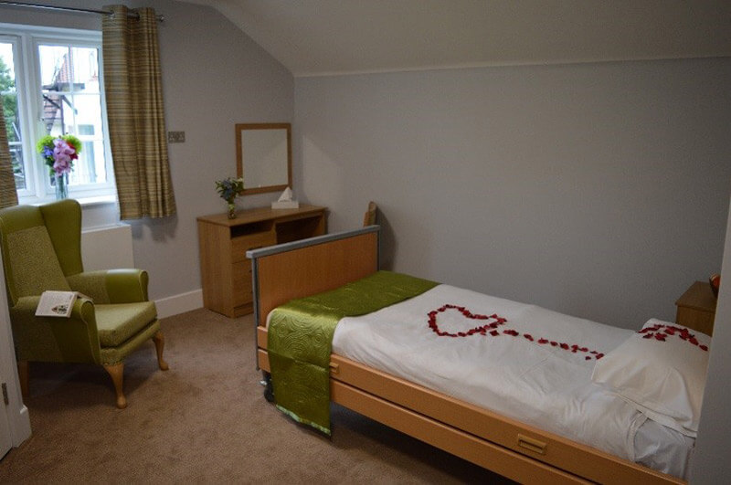 A bedroom with light wood bed frame and draw with white sheets, red petals and an olive green throw on the bed.