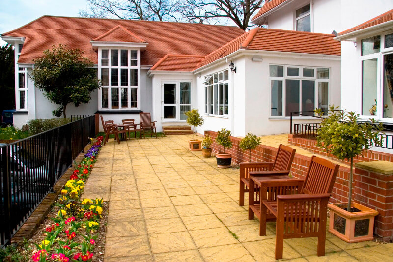 Stone patio area at the back of the care home with wooden bench seats and planted beds.