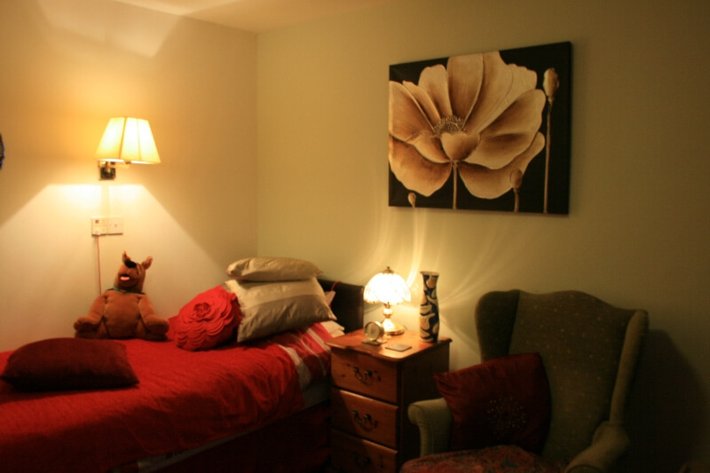 A bedroom with a lit lamps, with red duvet set and throw cushions at Marlborough House Care Home.