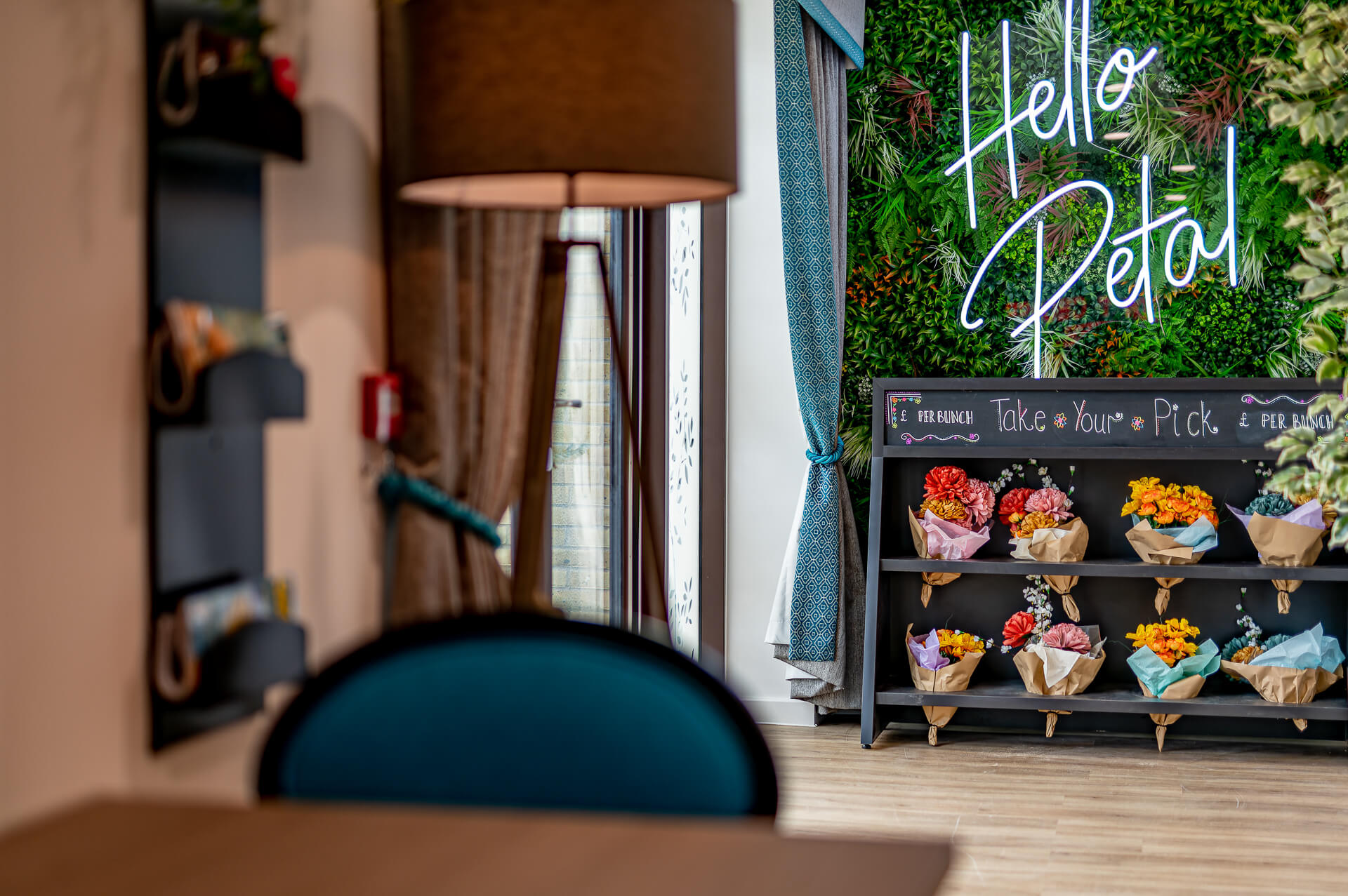 A flower stand in front of wall of green plants and a neon blue 'Hello Petal' sign at Surbitonian Gardens Care Home.
