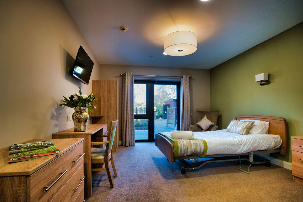 A bedroom with olive green feature wall, a bed, a table, a chest of drawers, a tv, a floor to ceiling window and some flowers.