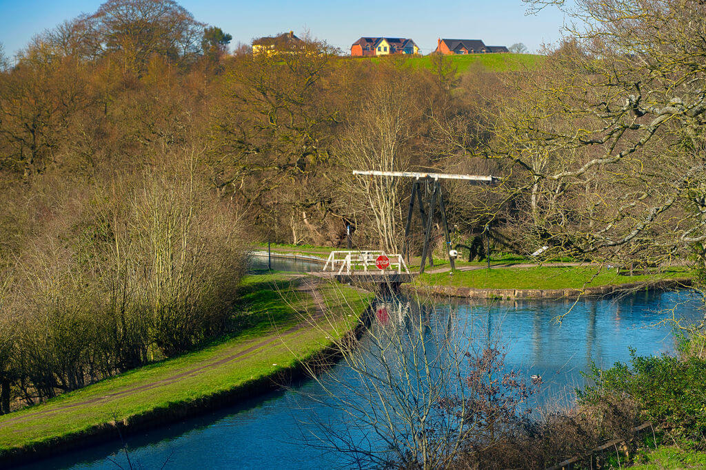A canal with a white bridge and lined with trees and grass verges.