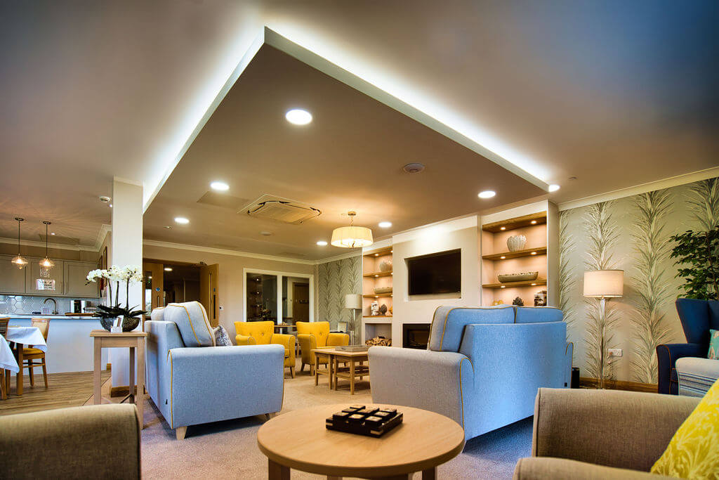 Tables, sofas and arm chairs in a communal lounge with TV mounted on the wall.