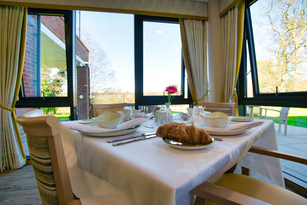 A table laid with a white table cloth, place settings and a plate with a croissant on.