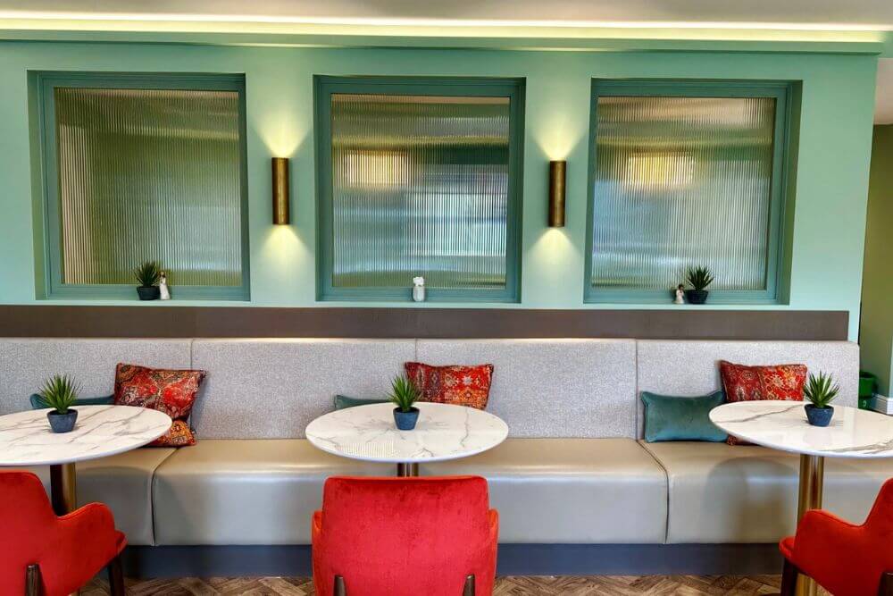 A grey booth bench with cushions, a round table and a red chair opposite the bench.