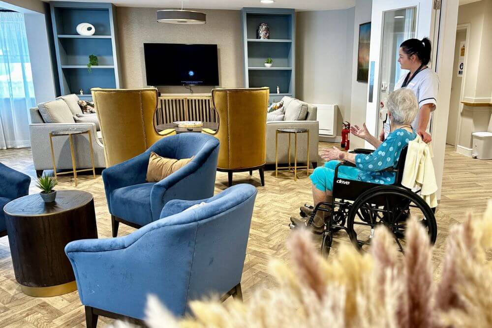 Sofas and armchairs in a lunge area with woman resident being wheeled in by woman carer.