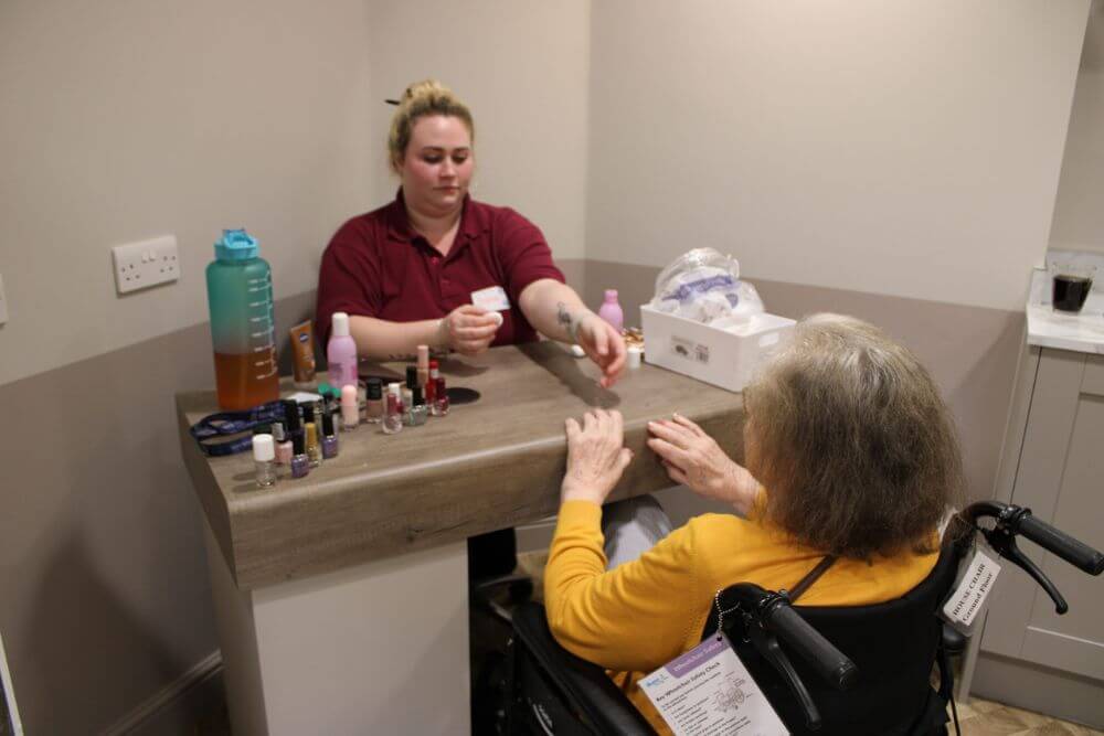 Woman sitting in her wheelchair having her nails painted by another woman.
