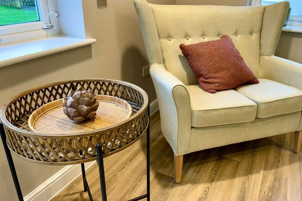 Pale yellow double arm chair next to a wicker, wood and metal round basket table.