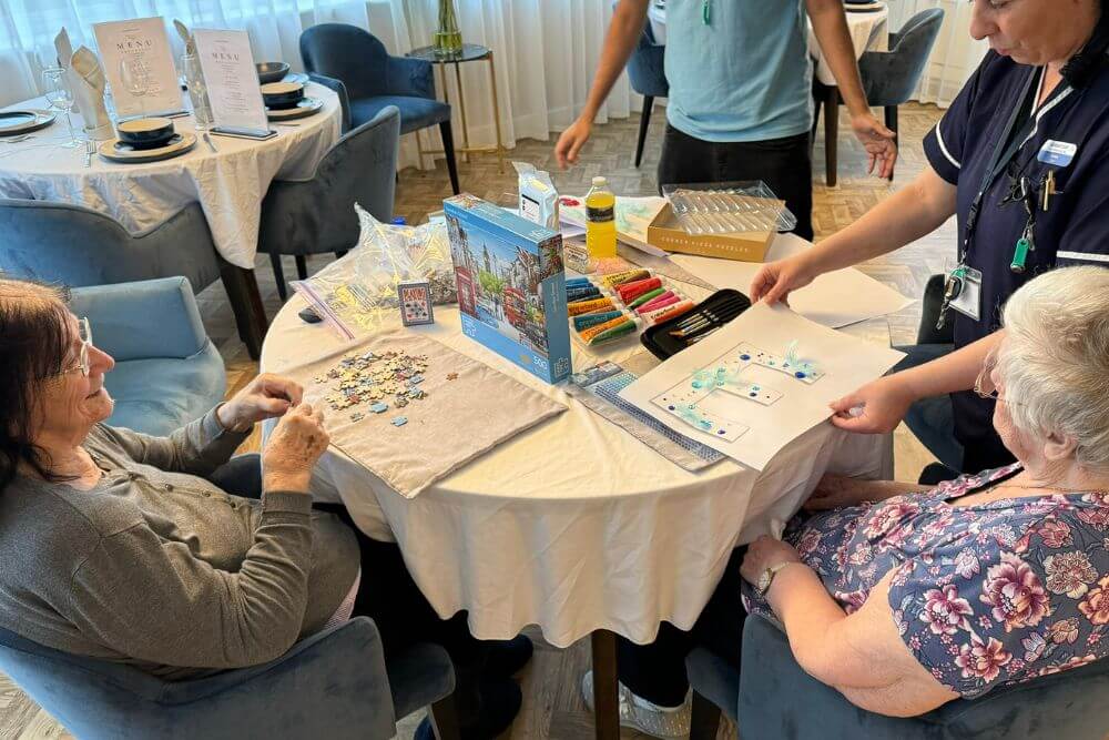 Woman doing a puzzle next to a woman doing a craft with paints on a round table.