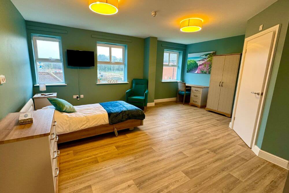 Large room with sage green walls, a single bed, double wardobe, desk and TV.